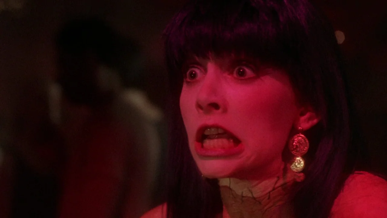 Frankenhooker (1990) movie still: closeup — A dark indoor space is bathed in intense red light. A young woman with dark hair stares with a wide-eyed…