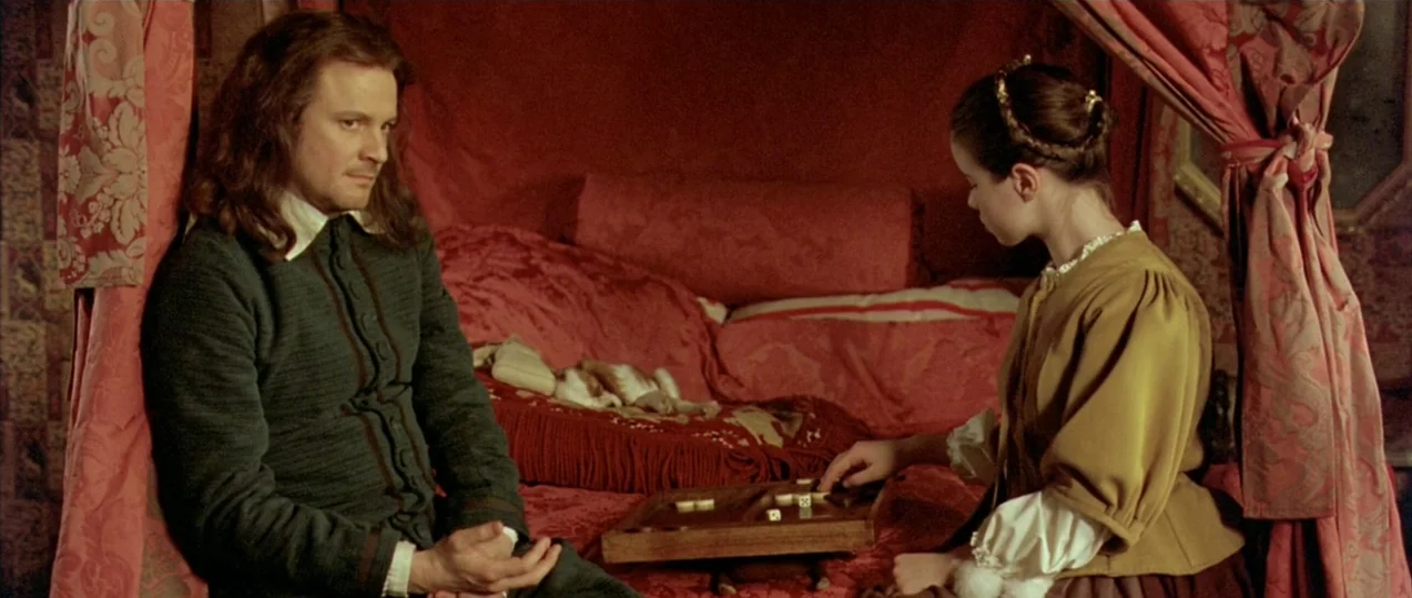 Girl with a Pearl Earring (2003) movie still: medium — A man and a woman sit together on a plush bed with red fabric drapes. They are focused on…