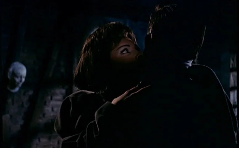 Mill of the Stone Women (1960) movie still: medium — A dimly lit room with stone walls and a suspended pale mask on the wall. A woman looks up…