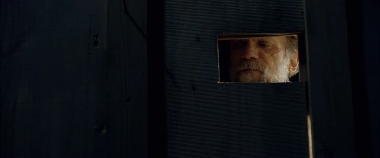 Ghosts of the Ozarks (2022) movie still: closeup — A close-up view shows a man looking through a small rectangular hole in a wooden wall. The dark…