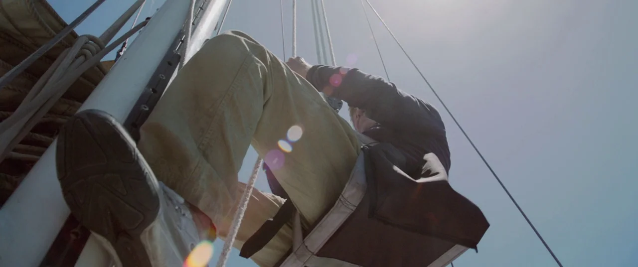 All Is Lost (2013) movie still: medium — The frame shows a person suspended by a harness against a white sailboat mast. Bright blue sky is visible…