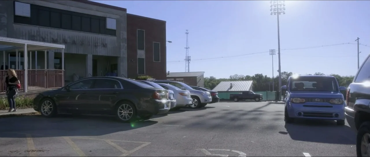 If You're Gone (2019) movie still: wide — A paved lot in front of a brick building during a sunny day. Multiple cars are parked in rows under a…