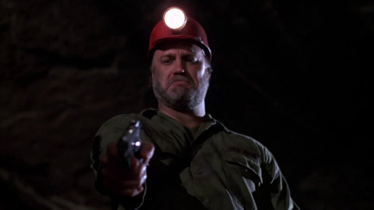 Day of the Dead (1985) movie still: medium — A dark subterranean space is lit only by a single bright headlamp. An armed man with a beard aims a…