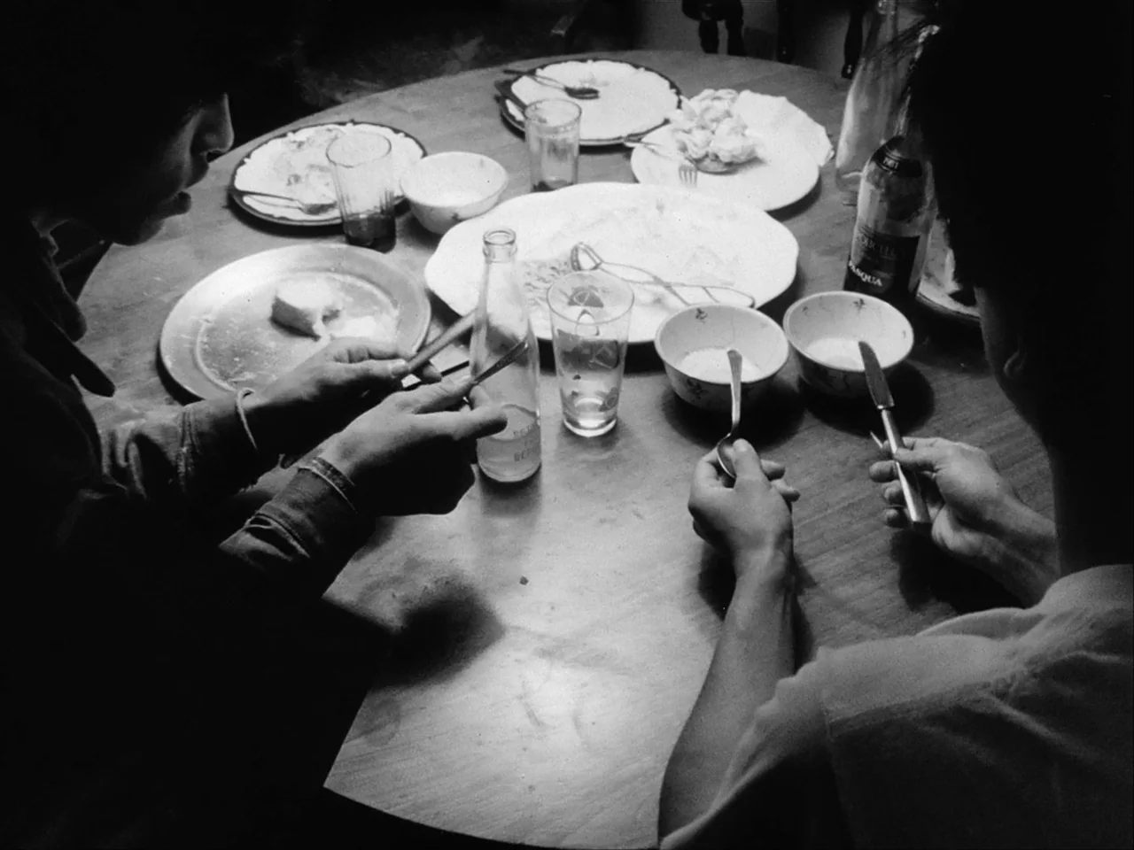 Mala Noche (1986) movie still: medium — A round wooden table is set with empty plates, bowls, and drinking glasses. Two people sit opposite each…