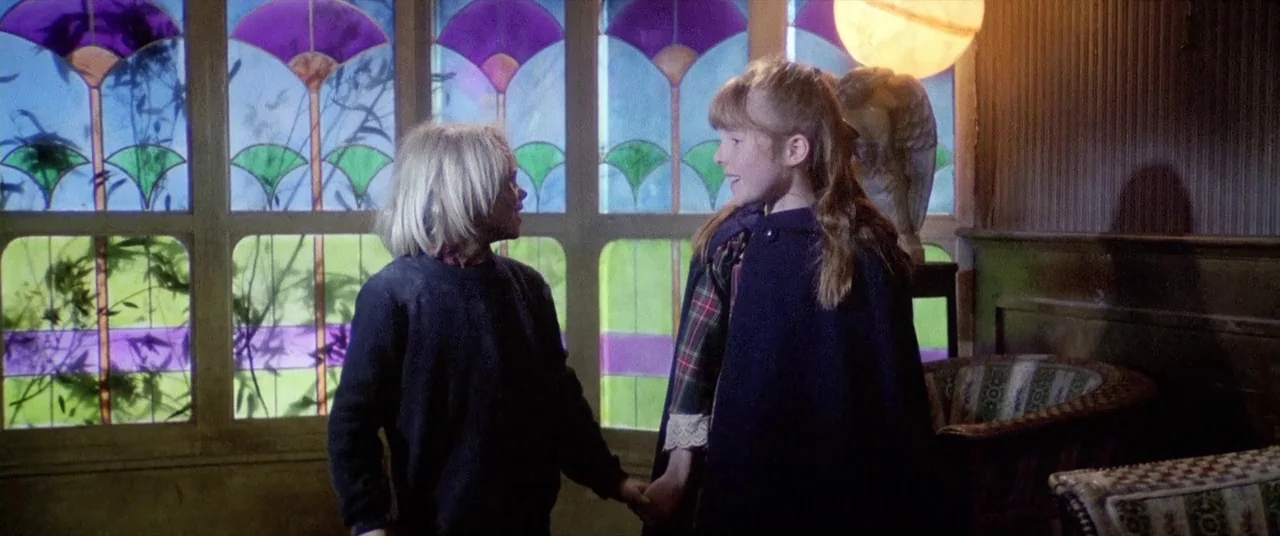 House By The Cemetery (1981) movie still: medium — Two children stand holding hands in a space filled with stained glass panels. The vibrant…