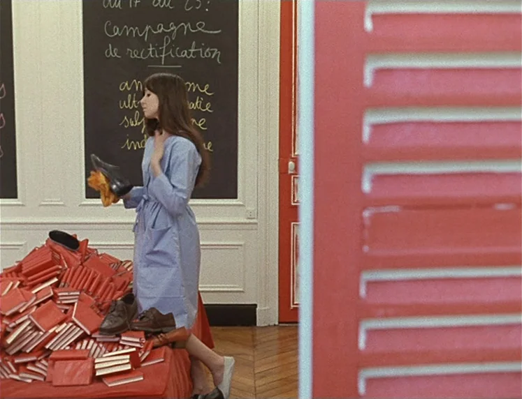 La Chinoise (1967) movie still: medium — A classroom with a tall chalkboard displaying white handwritten French text on the wall. A woman in a light…