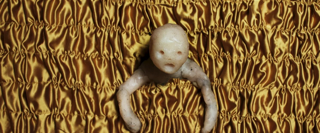 Stopmotion (2024) movie still: closeup — A pale, waxen figure with an open mouth and round eyes sits on gold satin. The fabric is tightly gathered…