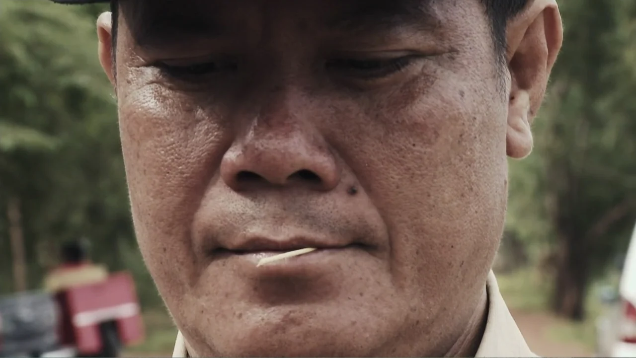 A Cambodian Spring (2018) movie still: closeup — A close-up of a middle-aged man's face in an outdoor setting with blurred greenery. The man has a…