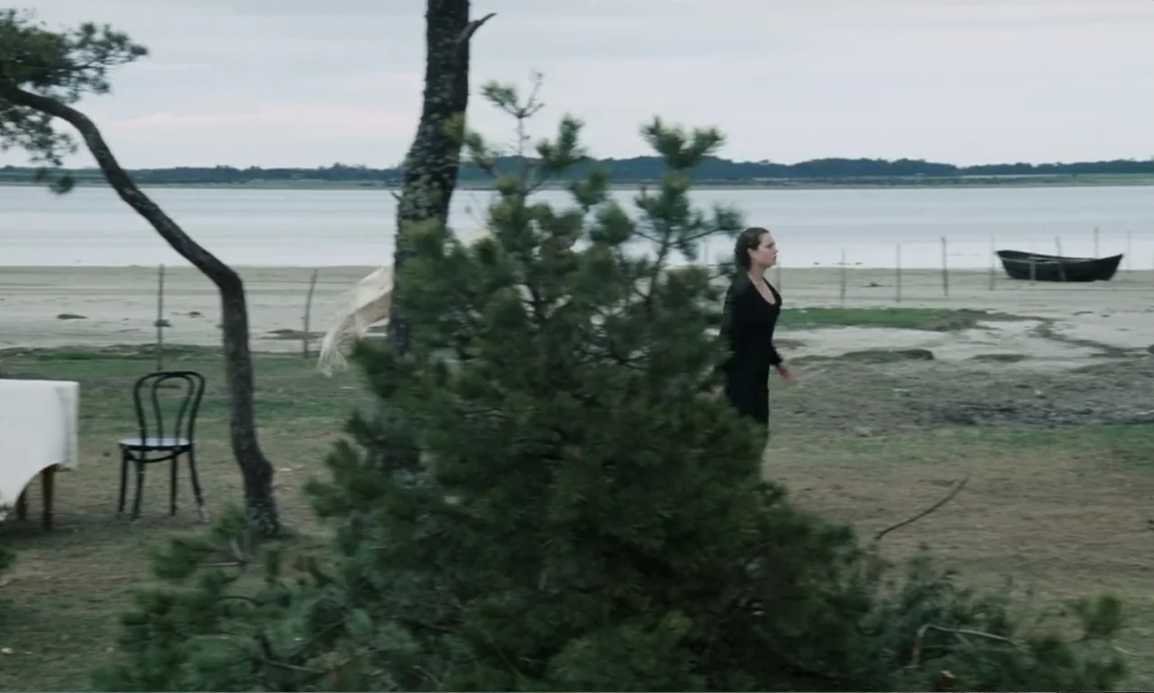 The Sacrifice (2015) movie still: wide — A quiet lakeshore landscape features a sparse grassy area with a single chair. A young woman in a black…