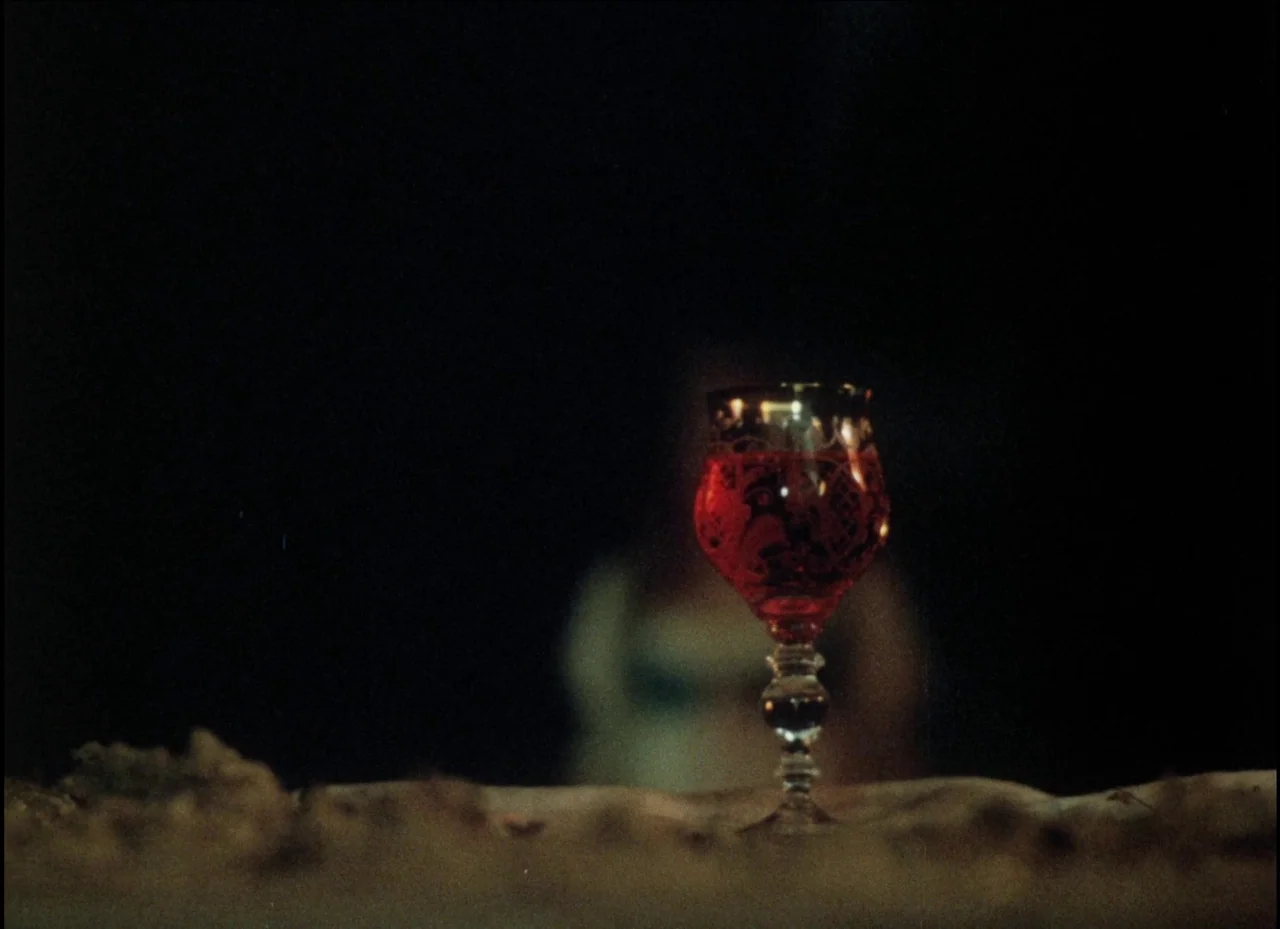 Beauty and the Beast (1979) movie still: closeup — A glass filled with red liquid sits on a table in a dark room. An out-of-focus figure is…