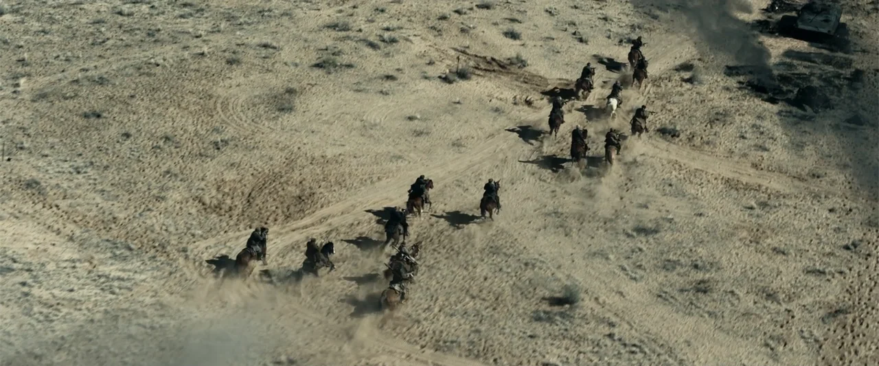 12 Strong (2018) movie still: establishing — A vast, arid desert landscape features a winding dirt track through light-colored sand. A group of…
