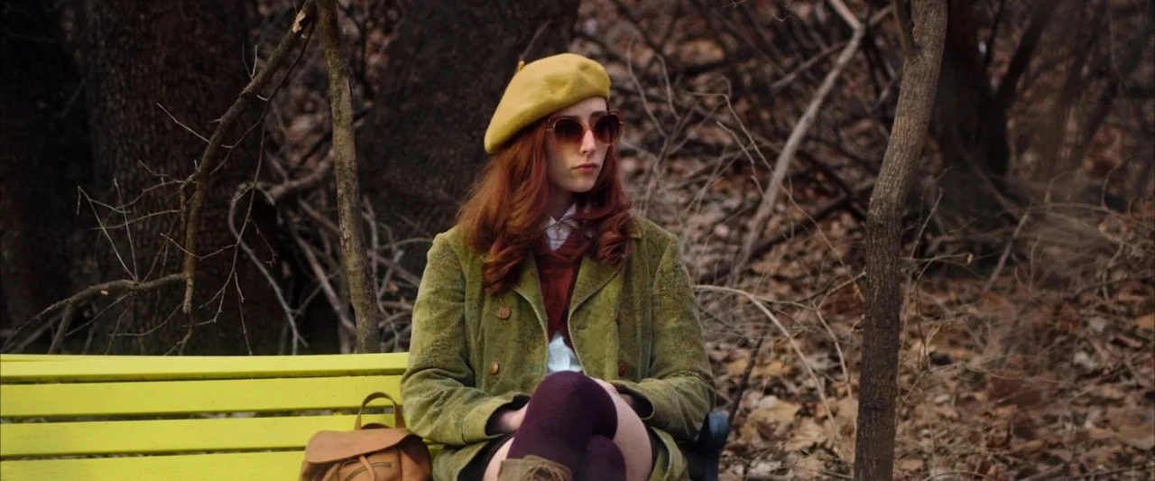 The Stylist (2020) movie still: medium — A woman sits on a bright yellow bench in a wooded area with bare trees. The woman wears a beret and green…