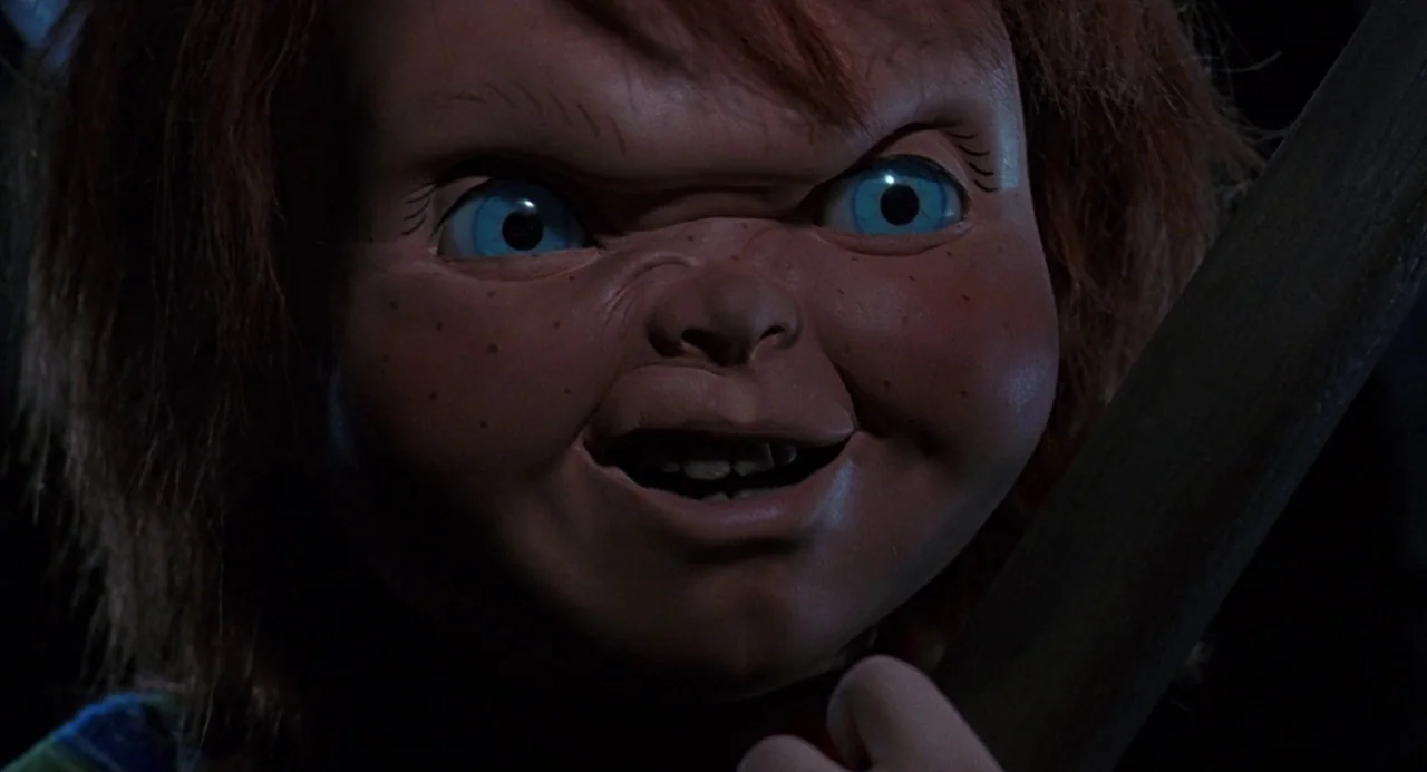 Child’s Play 2 (1990) movie still: closeup — A dark, out-of-focus background surrounds a close-up of a plastic doll face. The doll with bright blue…