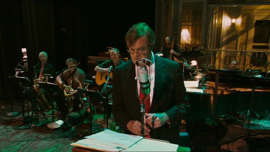 A Prairie Home Companion movie still: medium — A dimly lit broadcast studio set decorated with house silhouettes and warm floor lamps. A man in a…