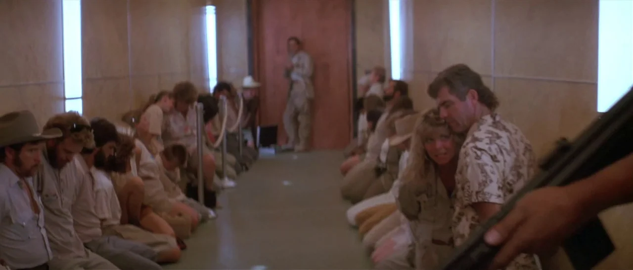 Universal Soldier (1992) movie still: wide — A group of people kneel in rows between beige walls in a brightly lit space. A man stands by a wooden…