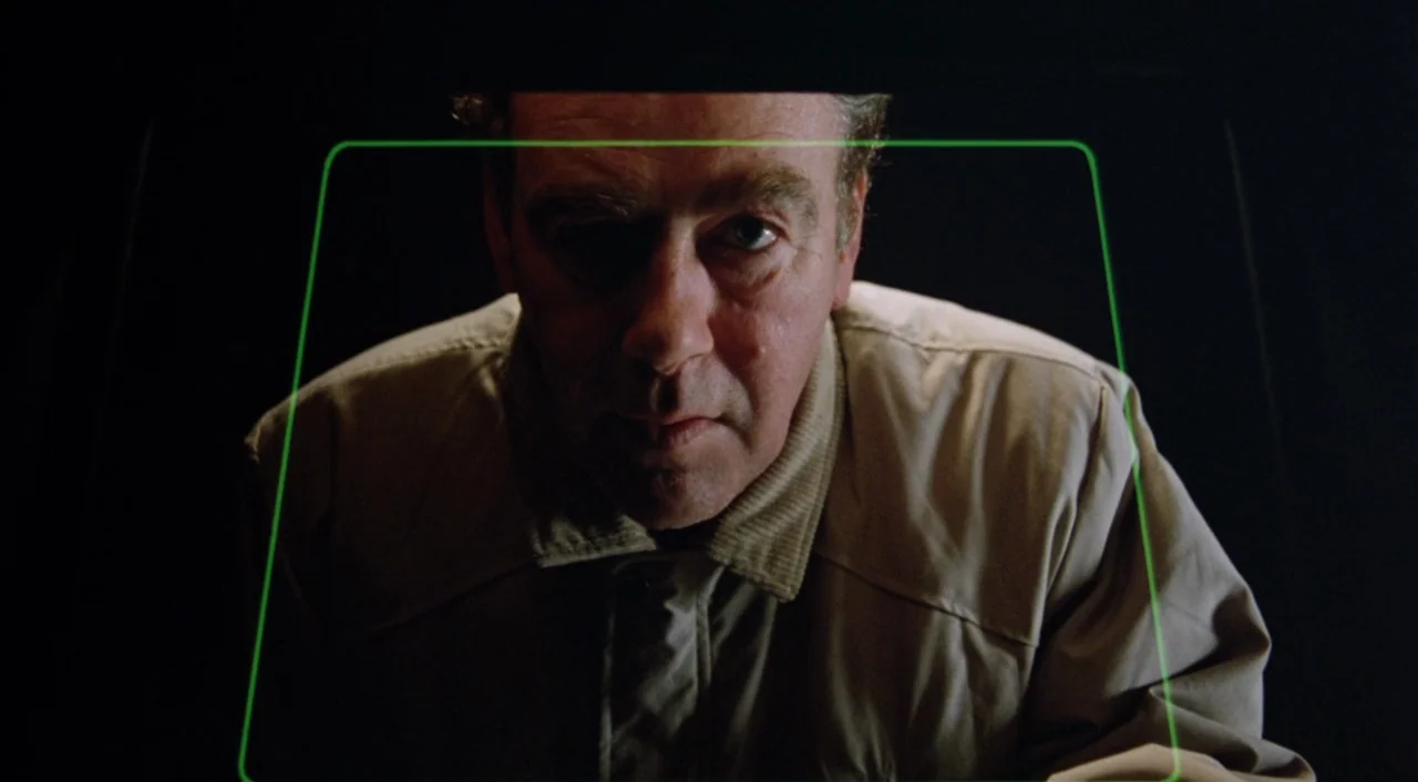 Shallow Grave (1994) movie still: closeup — A man looks directly into the camera lens within a dark, nondescript interior space. A luminous green…