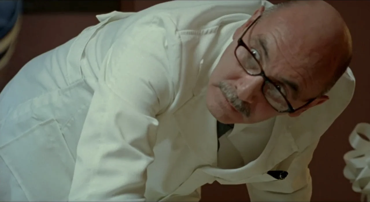 Sex, Love And Other Perversions II (2006) movie still: closeup — A man in a white lab coat leans forward in a sterile, dimly lit room. He looks…