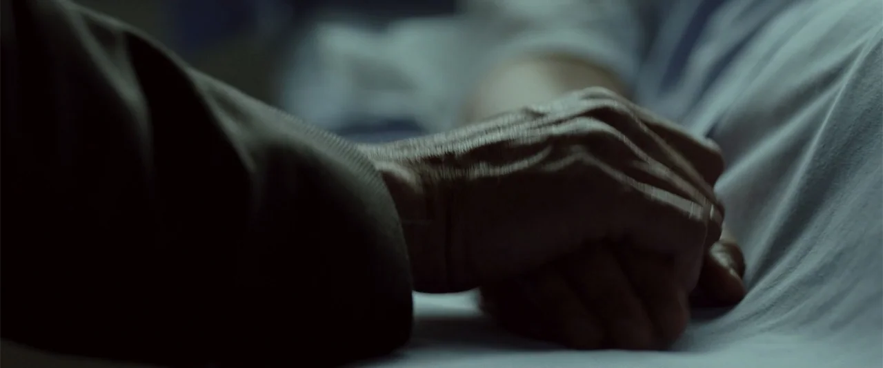 Anna (2013) movie still: closeup — A hospital room features white linens and a person wearing a textured sleeve. One hand rests on top of another…