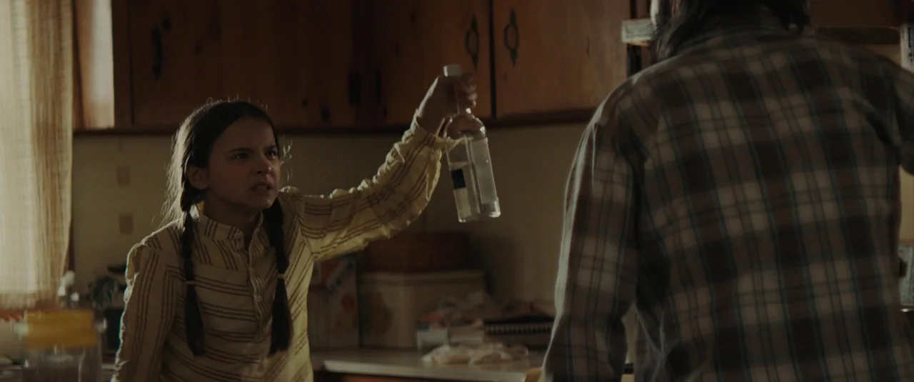 The Black Phone (2021) movie still: medium — A dimly lit kitchen features wooden cabinets and a cluttered countertop in the background. A young girl…