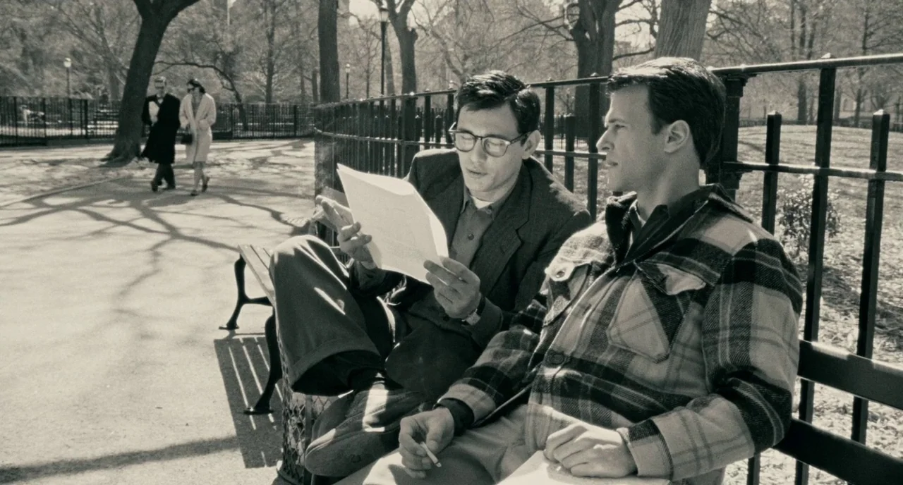 Howl (2010) movie still: medium — Two men sit on a metal park bench surrounded by leafless trees and paths. One man reads a document while the other…