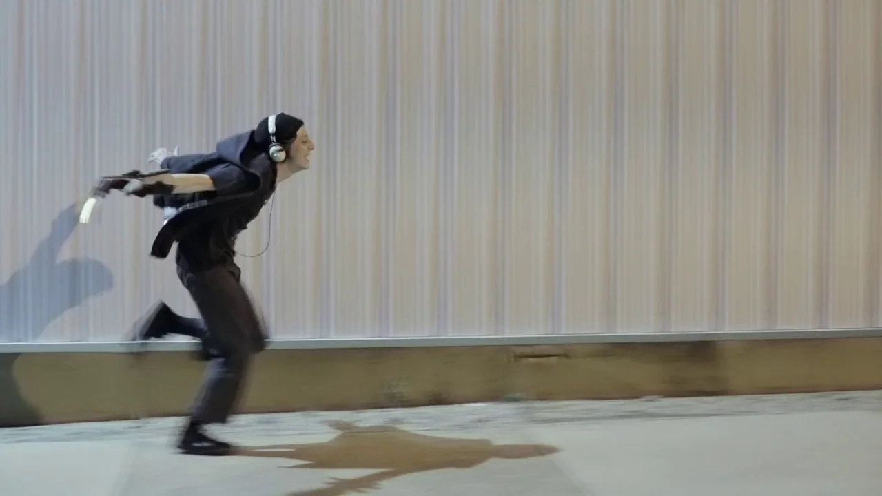 Buzzard (2014) movie still: wide — A person runs past a vertical corrugated metal wall on a paved surface. The subject is captured in motion blur…