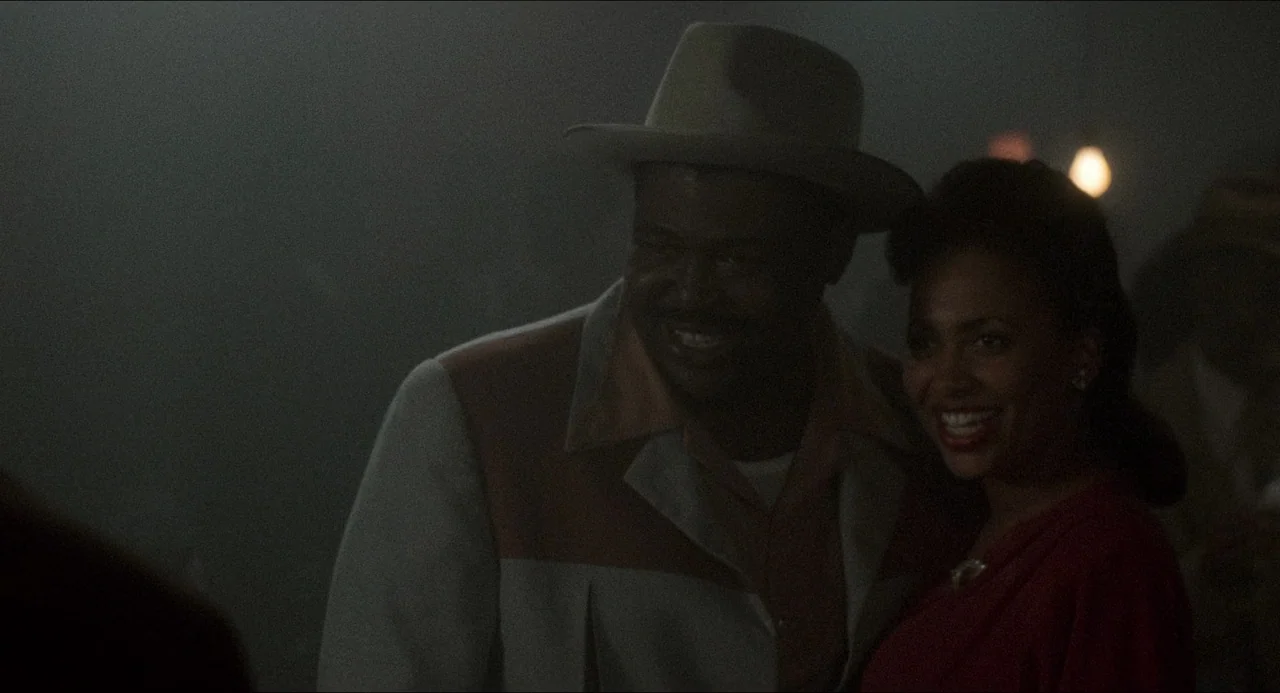 Devil In A Blue Dress (1995) movie still: medium — A dimly lit indoor space with hazy air and minimal visible background detail. A man in a hat and…