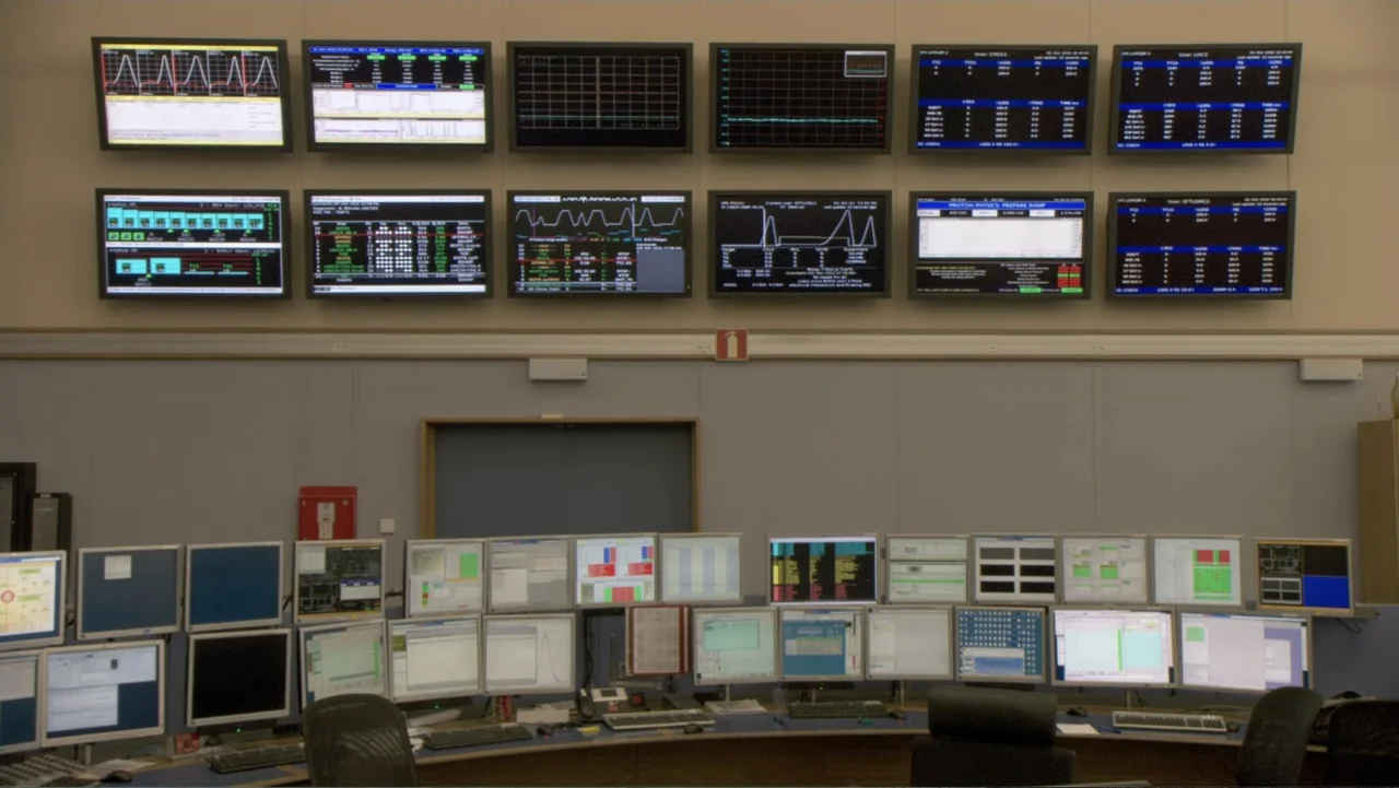CERN (2013) movie still: wide — A technical control room features rows of glowing computer monitors on walls and desks. The monitors display charts…