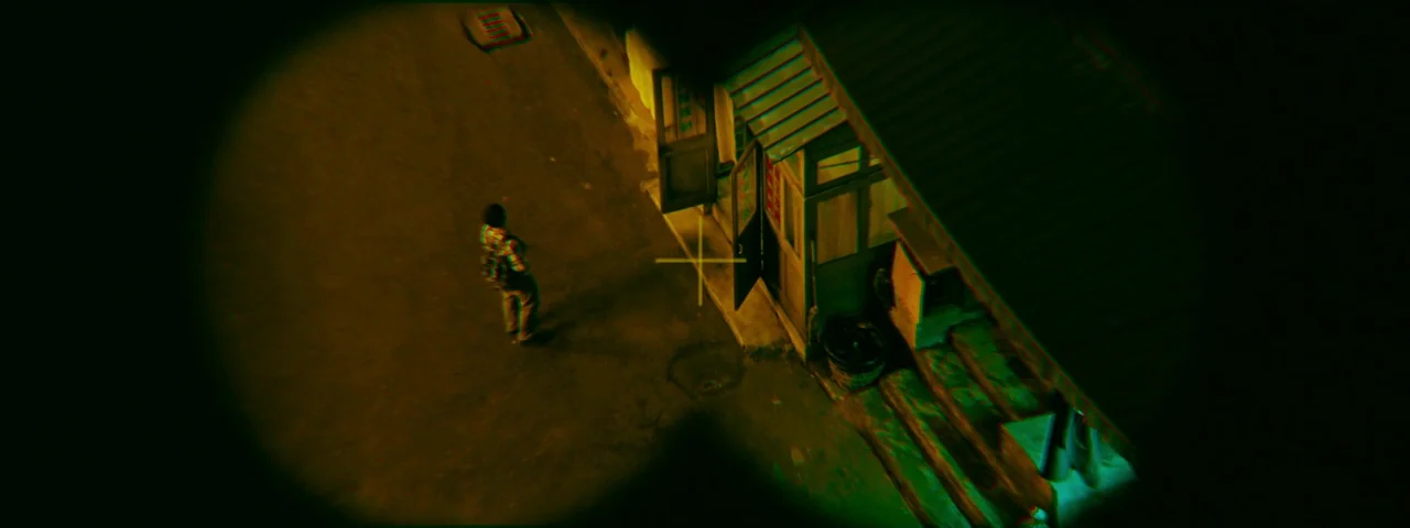 Under The Light (2023) movie still: wide — A dark night scene shows a building exterior viewed through a circular rifle scope. A lone figure in a…