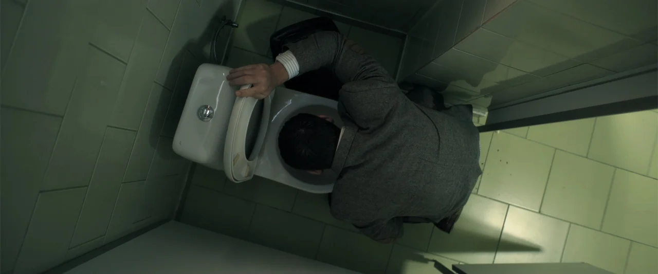 The Good Neighbor (2022) movie still: medium — A sterile tiled restroom stall features pale green wall tiles and matching floor tiles. A man in a…
