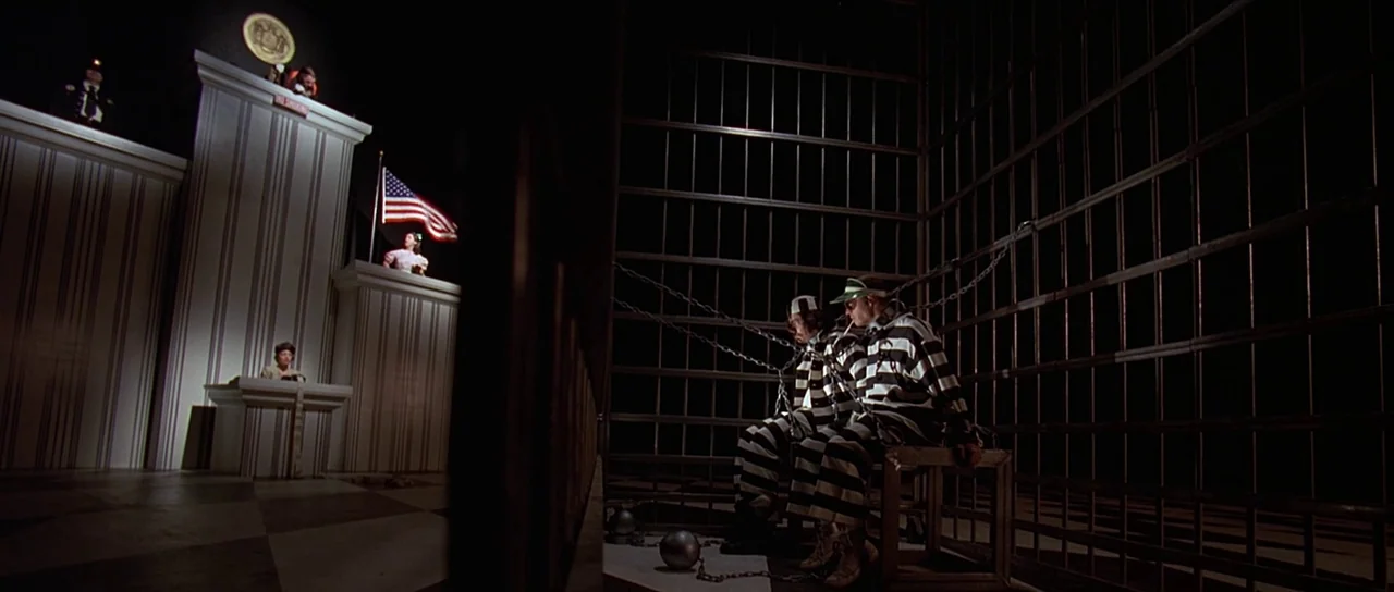Fear and Loathing In Las Vegas (1998) movie still: wide — A theatrical stage set mimics a prison scene with iron bars and chains. Two prisoners in…
