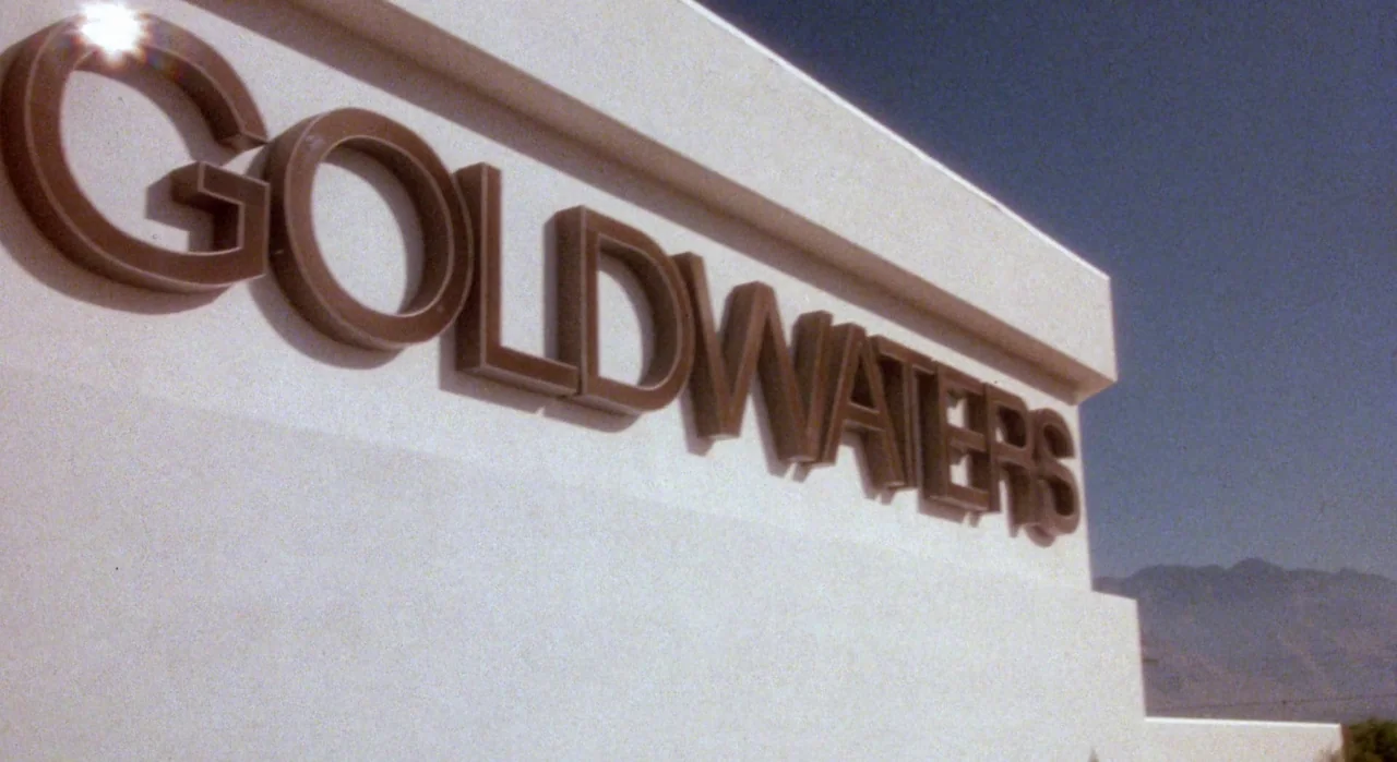 White Of The Eye (1987) movie still: establishing — A white concrete exterior wall displays a large, raised sign reading Goldwaters. The desert…