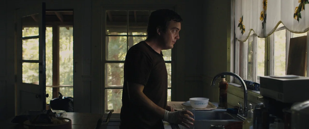 Blue Ruin (2013) movie still: medium — A dimly lit room with windows overlooking a treed area during the day. A man stands at the counter near the…