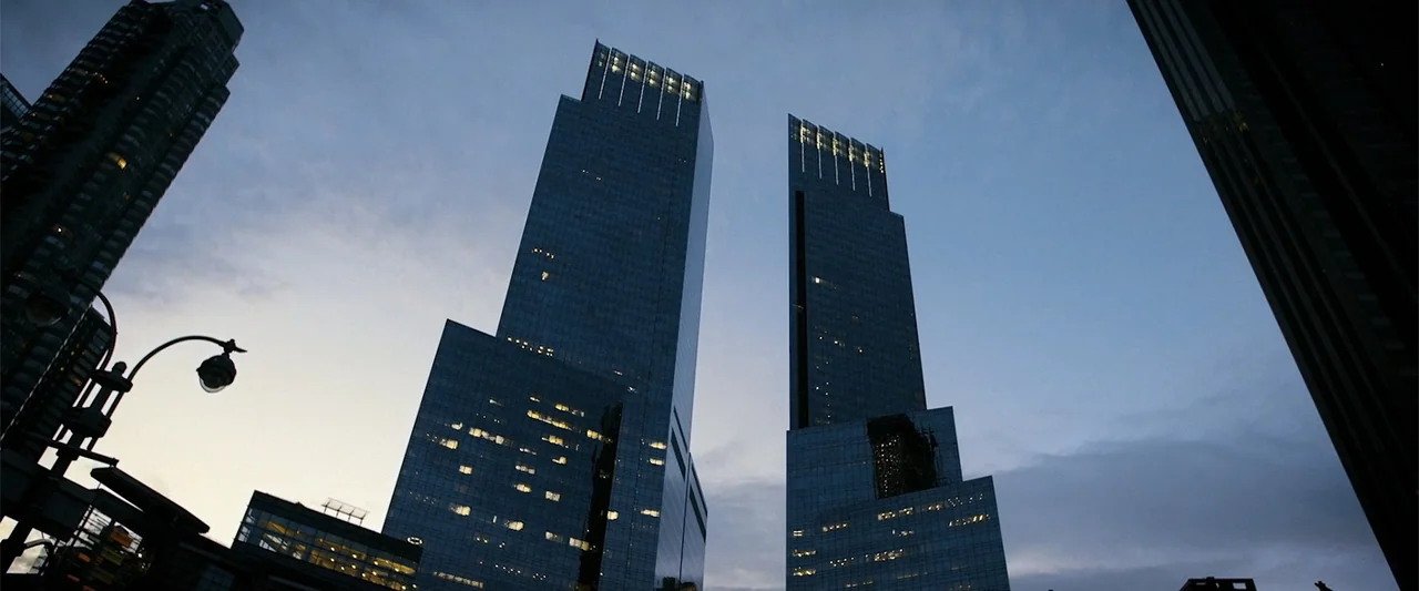 The Kleptocrats (2018) movie still: establishing — Two modern glass-paned skyscrapers rise toward a dusk sky in a city environment. The buildings…