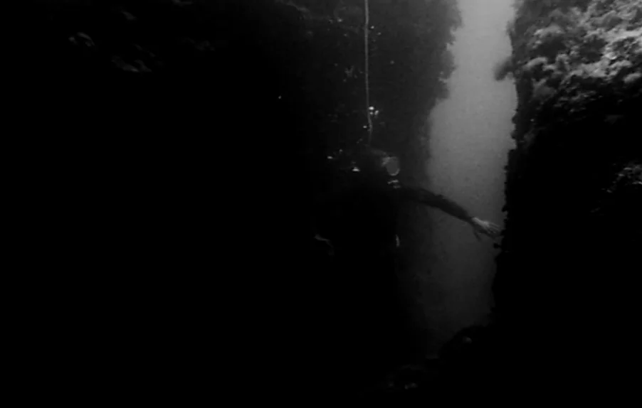 Caltiki, The Immortal Monster (1959) movie still: wide — An underwater passage is formed by dark, jagged rock formations. A lone scuba diver swims…