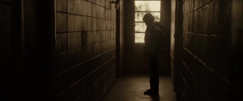 Let Me In (2010) movie still: fullbody — A long, dimly lit apartment hallway features exposed concrete block walls on both sides. A person stands in…