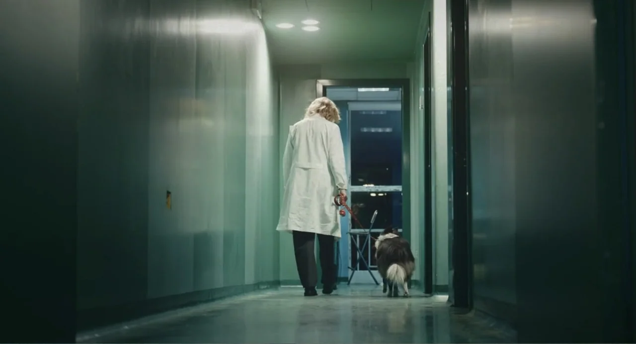 Elles (2018) movie still: fullbody — A long institutional hallway with smooth grey walls and clinical floor tile. A person in a white coat walks…