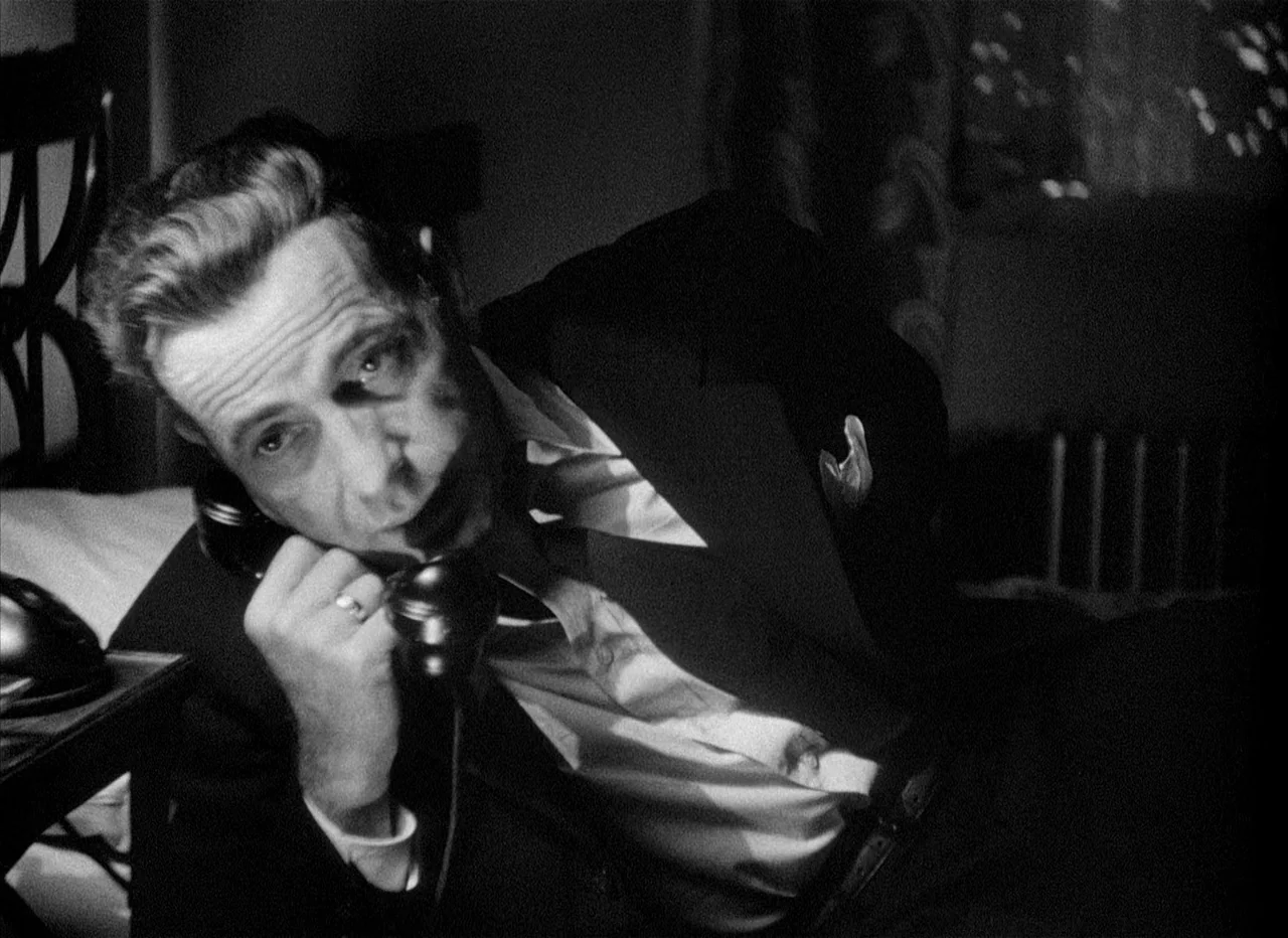Dead Reckoning (1946) movie still: closeup — A dimly lit room with a man reclining on a bed surface. The man holds a black rotary telephone receiver…