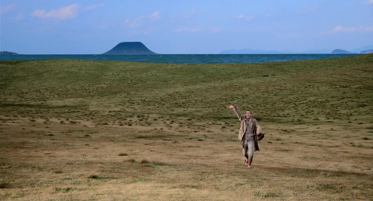 Hanagatami movie still: wide — A vast, rolling expanse of dry grass meets a calm, dark blue ocean. A man in tan clothing runs across the field while…
