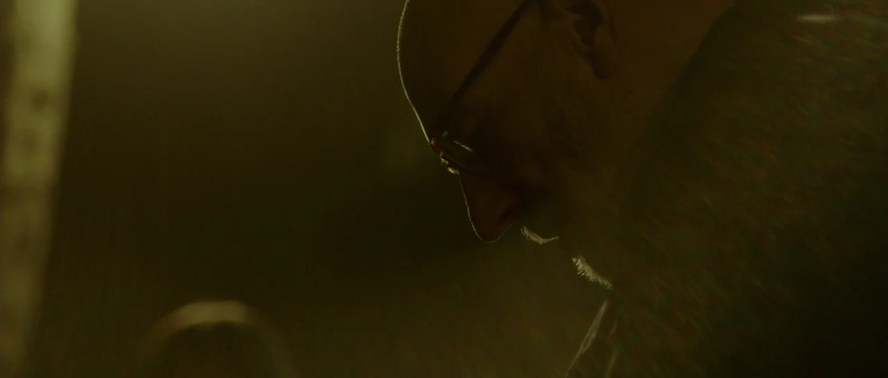 Green Room (2015) movie still: closeup — A dimly lit, indistinct space bathed in a monochromatic, dark-toned light. An older man with glasses stands…