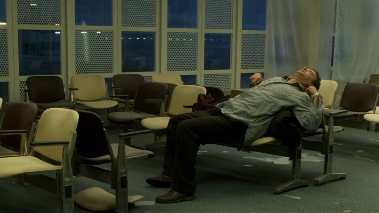 The Terminal (2004) movie still: medium — An empty airport terminal gate area features rows of tan and brown waiting chairs. A man in a striped…