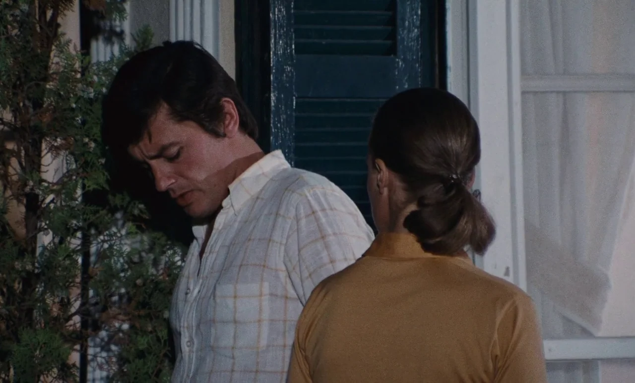 The Swimming Pool (1969) movie still: medium — A man and woman stand close together in front of a house door exterior. The man looks down while the…