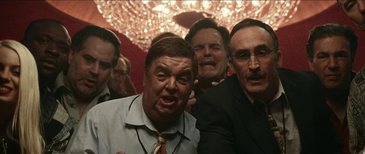 Bad Education (2019) movie still: medium — A crowded casino interior features a large crystal chandelier hanging above red walls. A diverse group of…