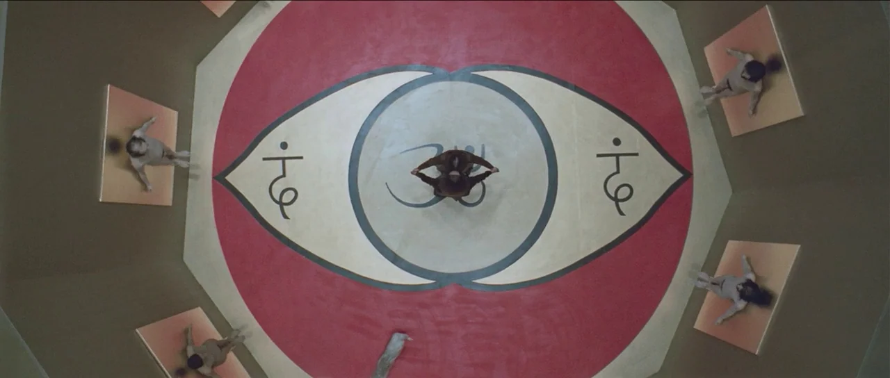 The Holy Mountain (1973) movie still: wide — A circular room features a large, symmetrical eye-shaped mural painted on the floor. Several figures…