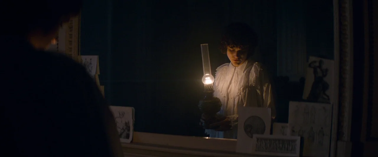 The Dancer movie still: medium — A dimly lit room at night features a large mirror framed by various sketches. A woman in a white nightgown holds an…