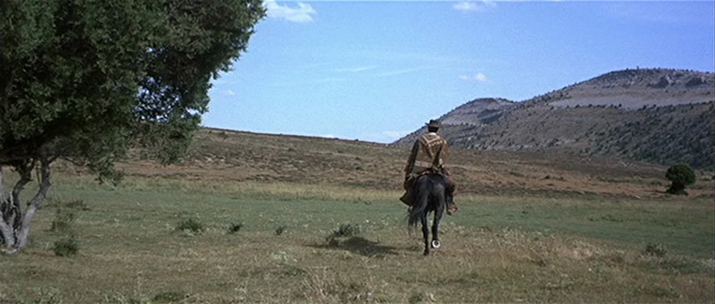 The Good, The Bad And The Ugly (1966) movie still: wide — A wide, open grassy meadow sits beneath a blue sky with rolling hills. A man on a horse…