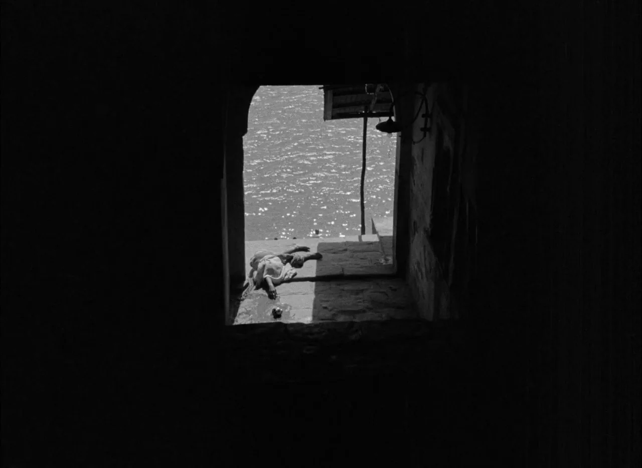 Aparajito (1956) movie still: wide — A dark stone threshold frames a sunlit waterfront scene with rippling water. A person rests motionless on the…