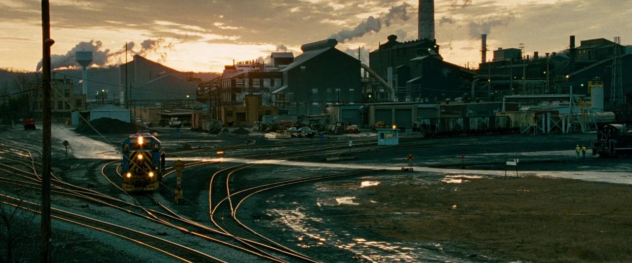 Unstoppable (2010) movie still: establishing — An industrial rail yard sits before a massive factory complex under a cloudy sunset sky. A blue and…