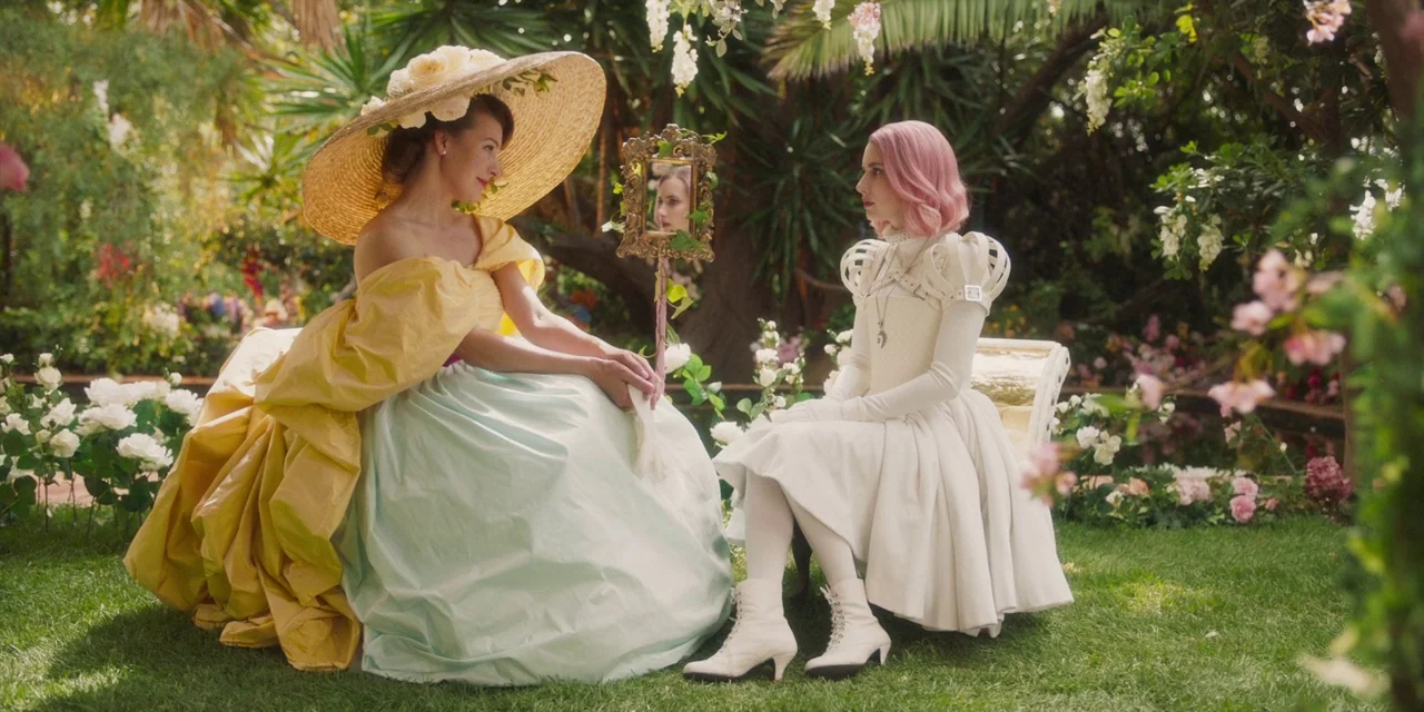 Paradise Hills (2019) movie still: medium — A lush garden with abundant white flowers and large tropical leaves surrounds two seated women. The…