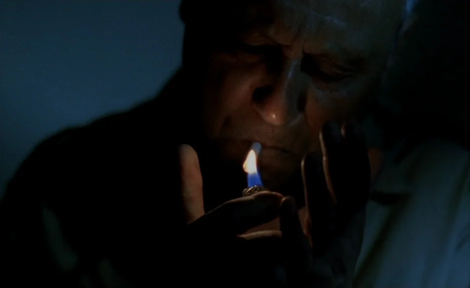 Beau Travail (1999) movie still: closeup — A dark indoor space with minimal ambient light. An elderly man holds a lit cigarette lighter near his face.