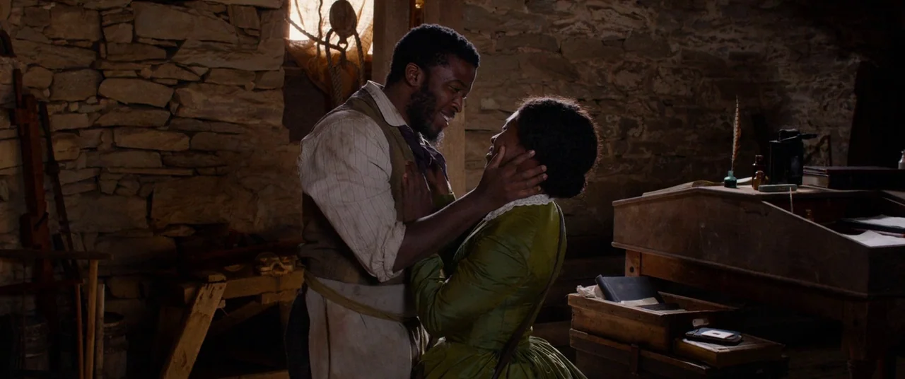 Harriet movie still: medium — A rustic workshop features unfinished stone walls and wooden construction tools. Two people stand facing each other…